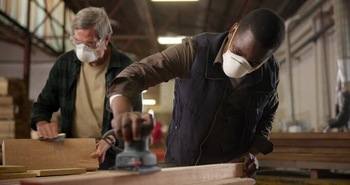 Men, carpenter and sanding woodwork in factory with machine for production process and manufacturing. Handyman, teamwork and mask with sandpaper tools in workshop for diy restoration and remodeling
