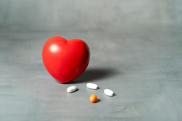 A red heart stress ball and various pills arranged on a smooth gray textured surface.