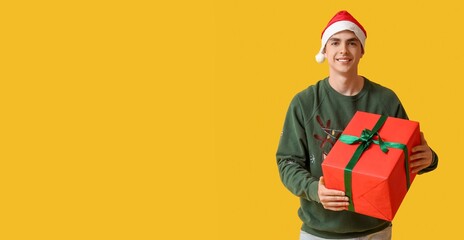 Happy young man in Christmas sweater and Santa hat holding gift box on yellow background with space for text