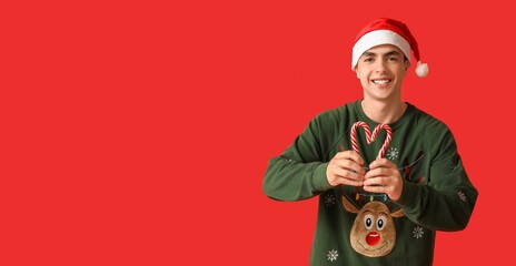 Happy young man in Christmas sweater and Santa hat holding candy canes on red background with space for text