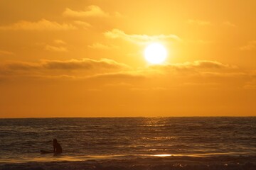 Surfer sitting on surfboard in ocean silhouette during golden hour sunset in Pacific beach, San Diego, California