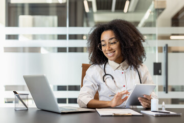 Confident female doctor using tablet and laptop in modern office setting, showcasing technology in...
