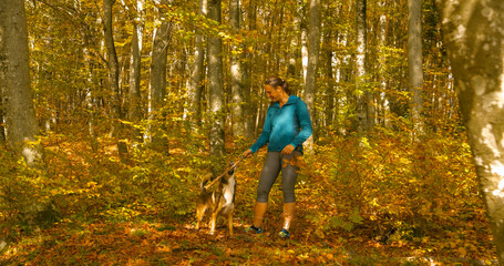 Woman in jacket playfully engages in a game with her dog, using a stick as they enjoy a sunny...
