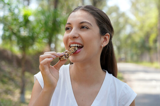 Beautiful young woman biting a nuts cereal energy bar in city park