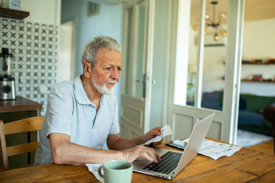 Elderly man managing bills and finances on laptop in kitchen
