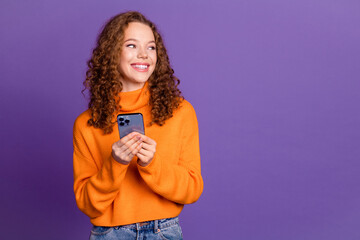 Portrait of pretty young girl hold phone look empty space wear orange sweater isolated on purple...