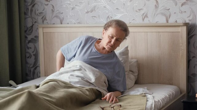 An elderly woman, weakened by illness, lying in bed, reaches for her glasses and falls back onto her pillow.
