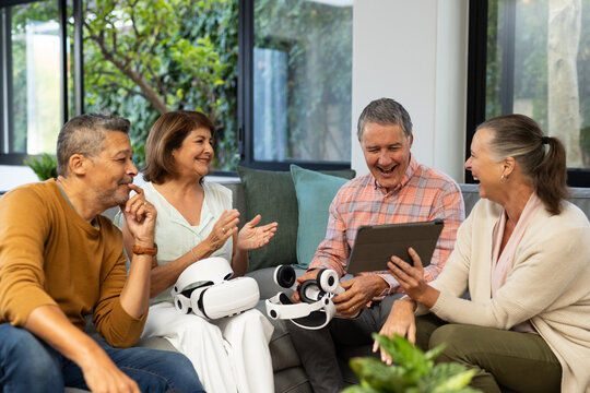 Using tablet, senior diverse friends laughing and discussing VR headsets in living room, at home