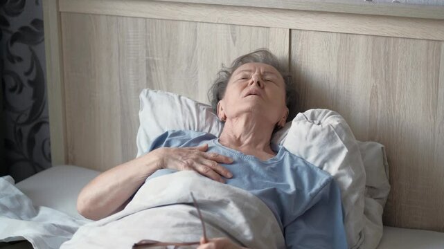 An elderly sick woman is breathing heavily, lying on the bed feeling a heaviness in her chest