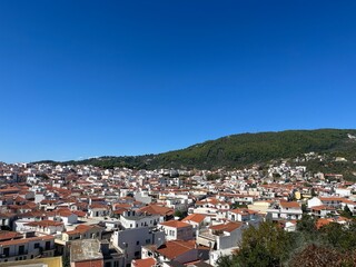 Fototapeta premium Panoramic View of Skiathos Town Greece – Scenic coastal hillside with white buildings and terracotta rooftops overlooking the Aegean Sea under clear blue skies