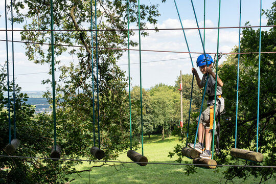 Young boy balancing on a rope bridge in an outdoor adventure park wearing a blue helmet and harness. Concept of outdoor activities, physical challenges, and adventure sports for children on a sunny