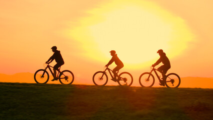 Fototapeta premium AERIAL, LENS FLARE, SILHOUETTE: Cinematic drone shot of a group of mountain bikers riding along a verdant hilltop at stunning orange sunset. Scenic view of friends riding electric bikes at sunrise.