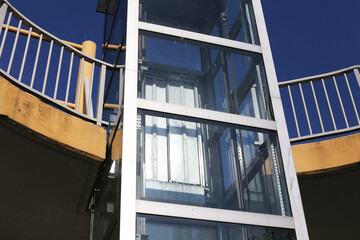 Glass elevator shaft against the sky background. Urban promenade with a functional elevator. Dirty glass panels from frequent use. Cityscape view with a modern, grunge weathered lift.