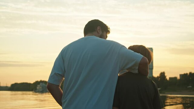 A young father provides support for his son. He embraces him, and together they observe the beautiful sunset over the lake.