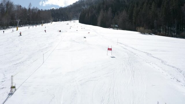 Ski slope under a clear sky with a racer navigating through the course marked by poles. Forested mountains line the background while other skiers make their way downhill.