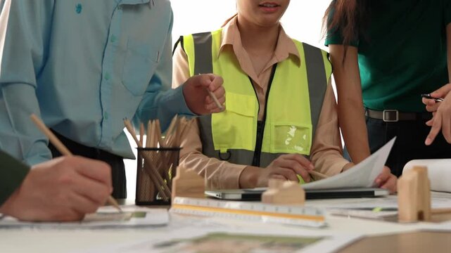 Smart architect and safety engineer discussion about building blueprint on table with architectural document, measurement tools and wooden block scatter around at modern office. Closeup. Delineation.