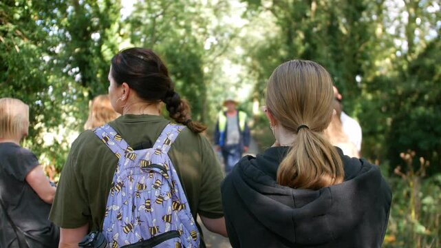 A group of people is walking along a tree-lined path, listening to a guide who is speaking ahead of them. The image captures an outdoor educational activity, such as a nature walk or guided tour