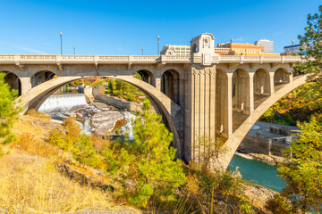 Fototapeta premium The Monroe Street Bridge, with the Lower Falls visible along the Spokane River and the downtown Riverfront Park skyline behind, on the Centennial Trail in Spokane, Washington.