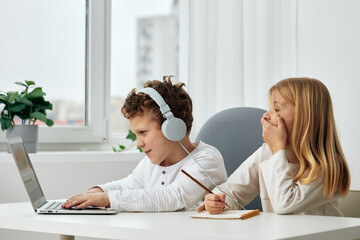 Fototapeta premium Happy children studying together online at home, using laptops and headphones The brother and sister are sitting at a table in their living room, concentrating on their elearning lessons The room is