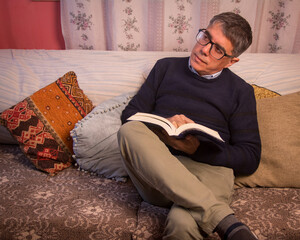 Fototapeta premium Adult man, wearing glasses, sitting on the armchair, thoughtfully reading a book