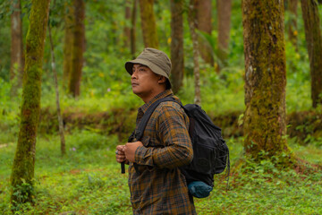 Half-body close-up of an adventurer in a dense and green pine forest. An Asian male traveler in a flannel shirt, bucket hat and backpack poses against a backdrop of dense pine trees.