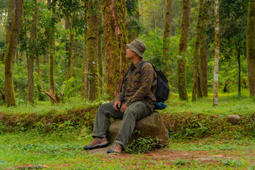 Fototapeta premium An adventurer is sitting on a large rock in a dense pine forest. An Asian man in a flannel shirt and bucket hat is sitting and resting while enjoying the tranquility of the beautiful nature.