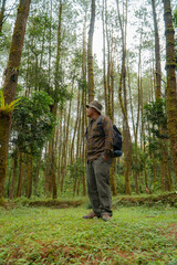 Asian man in adventure outfit in dense and green pine forest area. An adventurer in a flannel shirt, bucket hat, and backpack poses against a backdrop of dense pine trees.