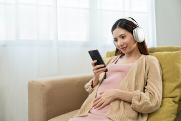 Happy pregnant young woman holding headphone on her womb at home, relaxed young asian mother let her fetal listening to music, family planning and pregnancy with insurance for child health.