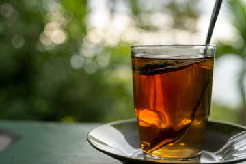 A glass of herbal tea with leaves in it, placed on a plate, with bokeh nature background. Natural and peaceful atmosphere for relaxing drinks outdoors.