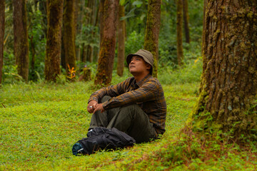 An adventurer is sitting on green grass in a lush pine forest area. Asian man in flannel shirt and bucket hat is sitting and resting while enjoying the tranquility of beautiful nature