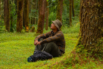 An adventurer is sitting on green grass in a lush pine forest area. Asian man in flannel shirt and bucket hat is sitting and resting while enjoying the tranquility of beautiful nature
