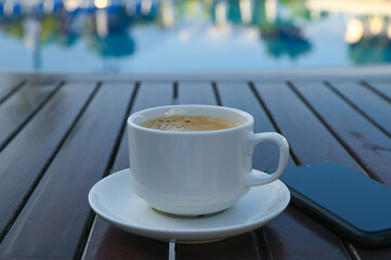 A cup of coffee with white foam on the surface on a wooden table. A phone in a black case lies nearby