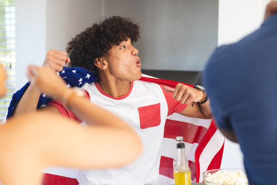 Cheering enthusiastically, man draped in American flag celebrating with diverse friends, at home