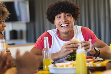 Eating burger, young man smiling and enjoying meal with diverse friends, at home