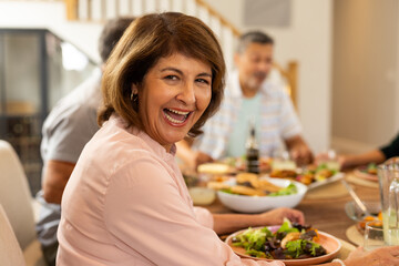 Smiling senior woman enjoying meal with diverse friends at dining table, sharing laughter, at home