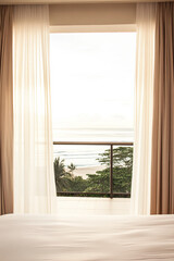 Fototapeta premium Coastal view through sheer curtains in a tranquil room with a balcony overlooking the ocean during early morning light