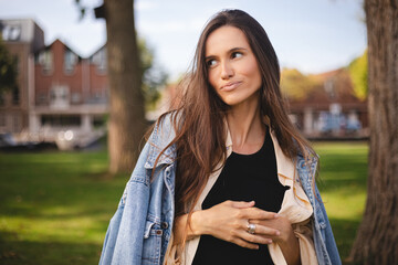 Close up portrait of beautiful confident woman laughing in nature, walk in park and look by side. Brunette woman look at side, enjoy of nature, amazement woman wear shirt, top and jeans jacket.