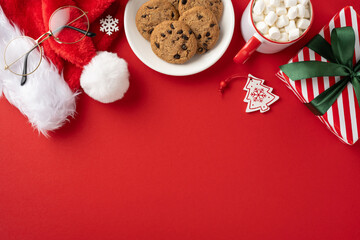 Top view of a Christmas scene featuring a Santa hat, cookies, hot cocoa with marshmallows, and a wrapped gift on a red background