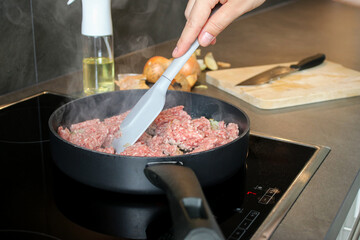 Close up a female hand stirring raw ground beef meat in a pan with a spatula, blurry background with onions and oil spray