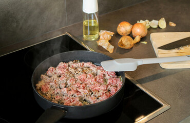 Close up raw ground beef meat frying in a pan with a spatulate, blurry background with onions and oil spray