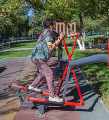 Obraz premium Elderly woman is doing exercise on a sports trainer ellipse. Workout in the yard. The concept of a happy old age and body positive. International Day of Older Persons and Grandparents Day