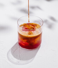 Espresso pouring into glass with red liquid and ice cubes on white background with shadows