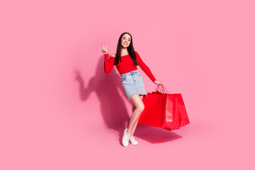 Full size photo of gorgeous young woman show v-sign hold bags isolated on pink color background