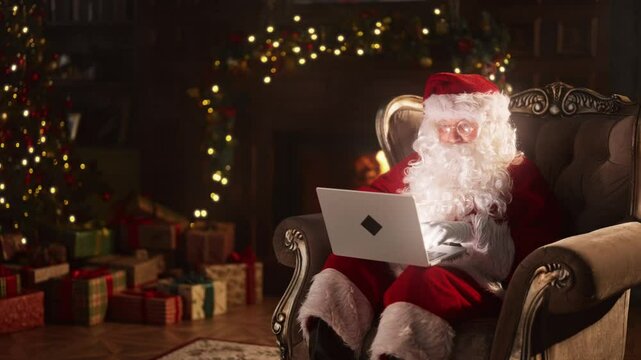 Santa Claus using modern laptop for communication and ordering gifts for kids. Old wizard with portable computer on laps sitting alone in cozy room with fireplace, preparation to Christmas festivities