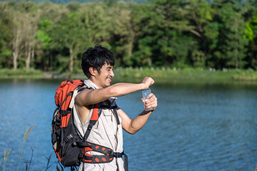 Thirsty hiker with backpack opening bottle and drinking water by scenic lake. Young Asian man.