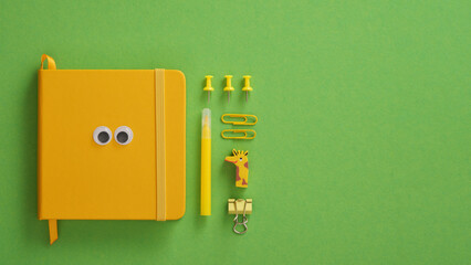 Yellow notepad with eyes and stationery items felt-tip pen, pins, eraser, paper clips on a green background. Space for your text.