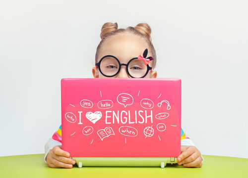 Beautiful cute little girl standing behind laptop with "I love English" phrase and drawings on cover.  Foreign language learning concept.