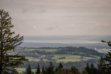 A scenic view of a Bavarian landscape with rolling hills, forests, fields, and a distant lake under a cloudy sky..