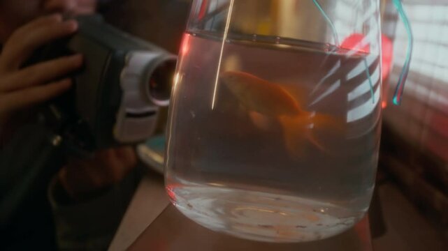 Closeup of young millennial puffy-haired boy making memories with pet goldfish swimming in aquarium, recording it on retro vhs camcorder, using outdated technology, nostalgic 90s