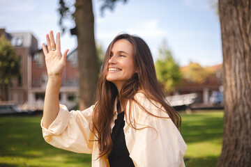 Young beautiful brunette woman with long hair congratulating her friend. Portrait of carefree friendly woman, smiling broadly while waving raised palm, greeting friend, meeting with mates walking park